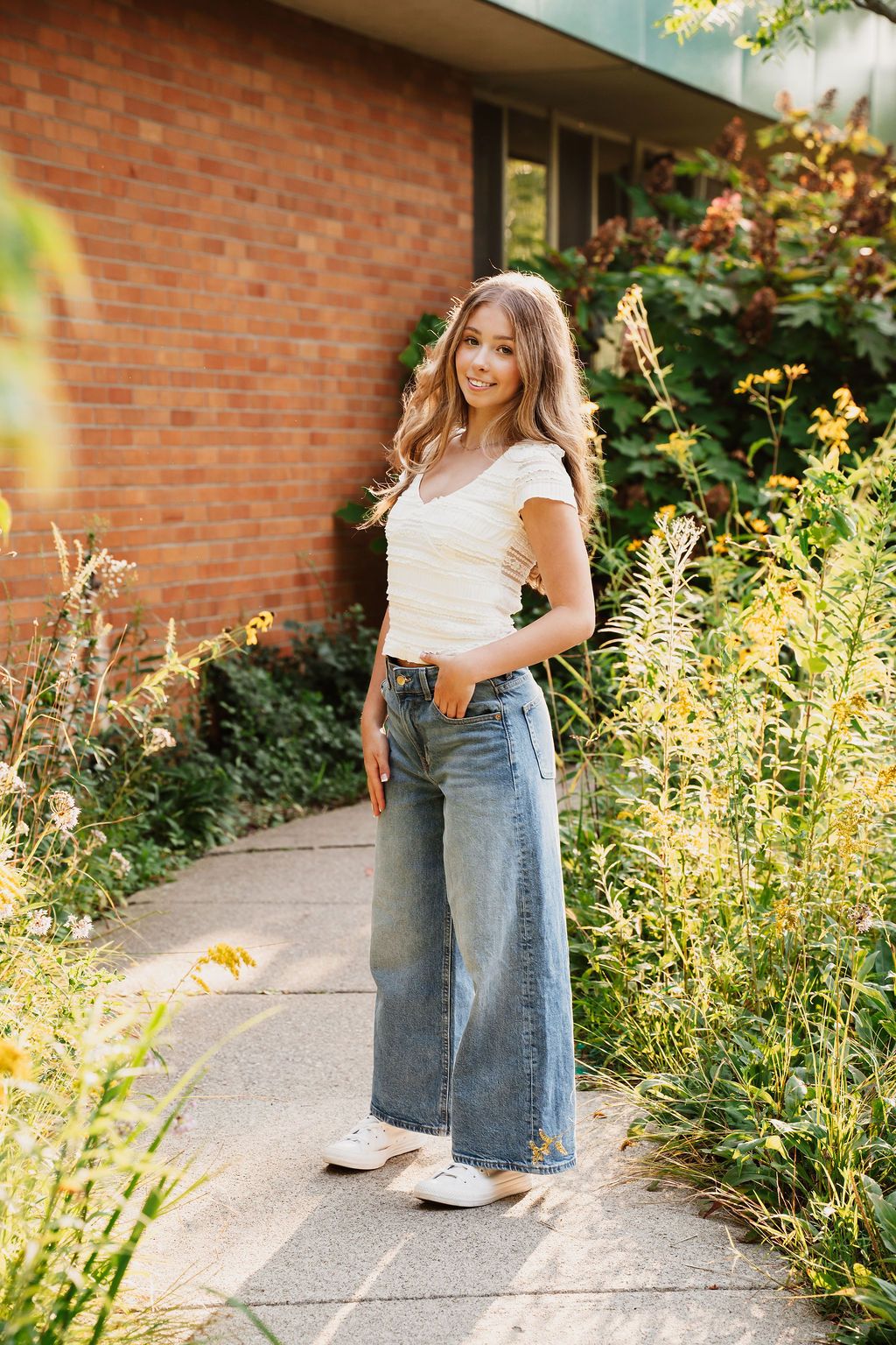 Summer portrait session featuring casual denim and white attire against a brick wall surrounded by golden wildflowers.