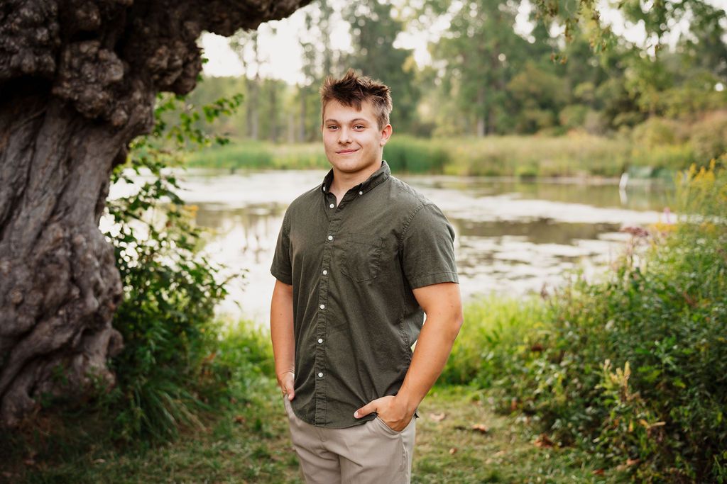 Young man in olive shirt and khaki pants smiling outdoors near a scenic pond surrounded by lush greenery.