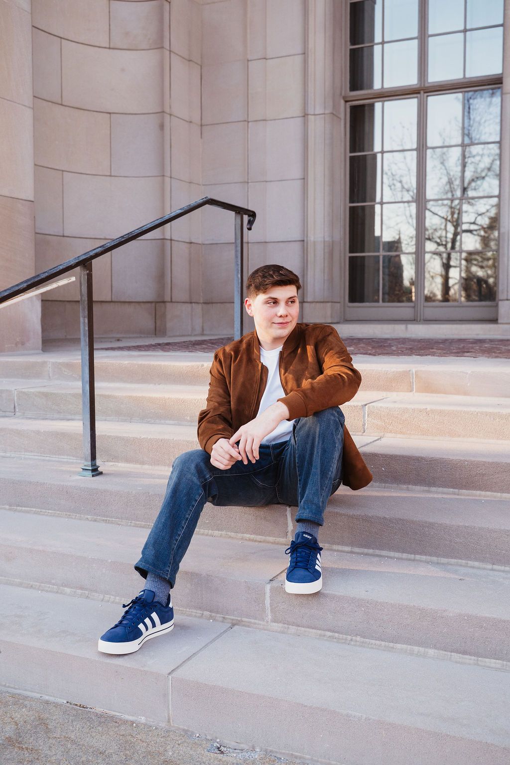 Young man in brown jacket and jeans sitting casually on stone steps outside a modern building.
