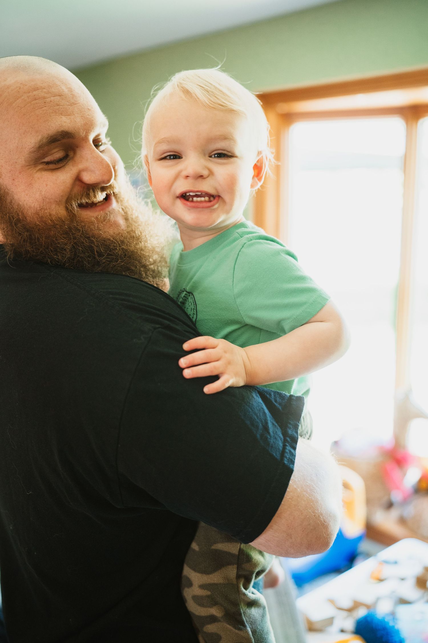 Bearded father lovingly holds smiling blonde toddler in green shirt, sharing a joyful moment indoors.