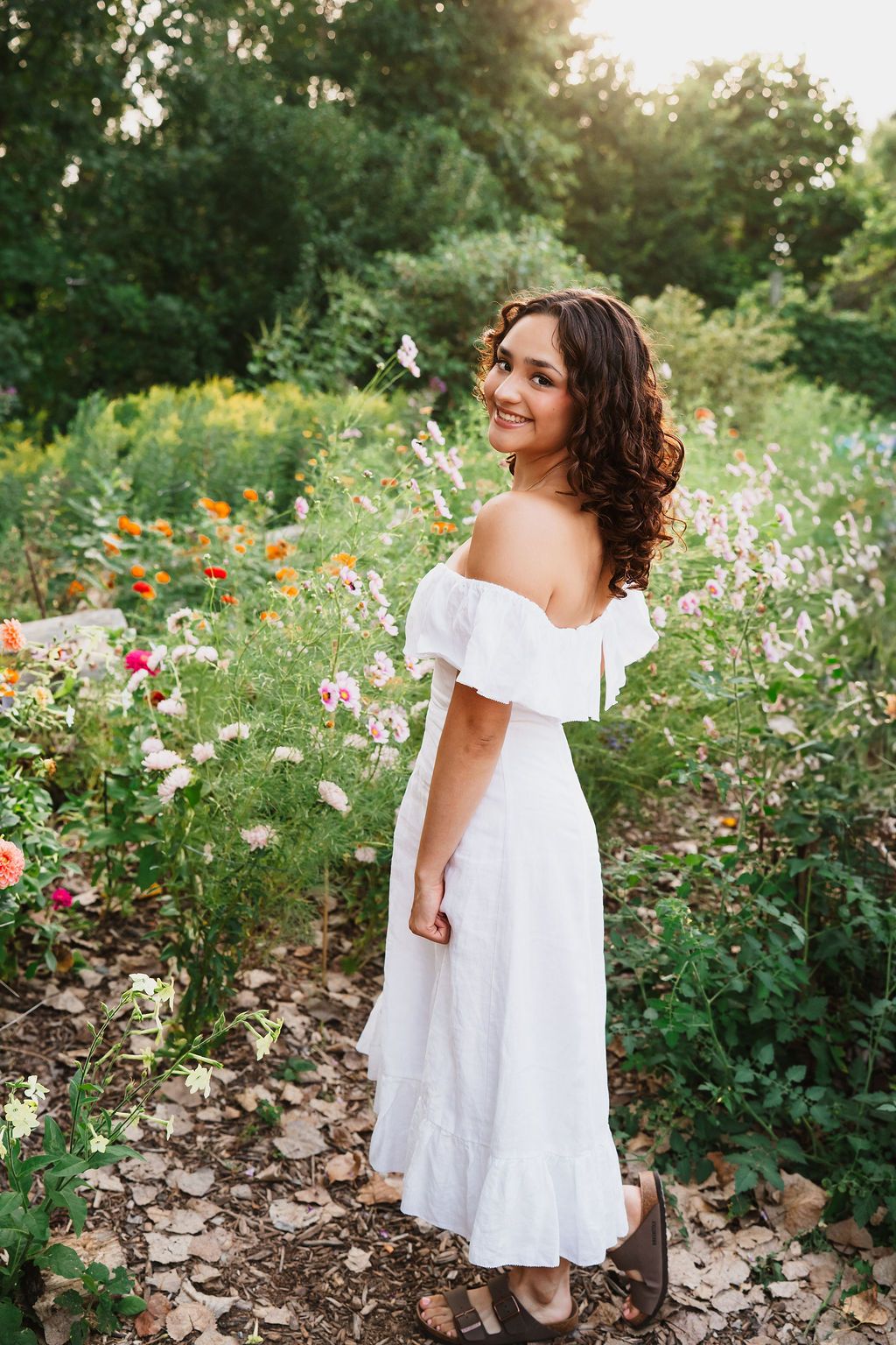 A person in a flowing white dress poses gracefully among vibrant wildflowers in a garden during golden hour.