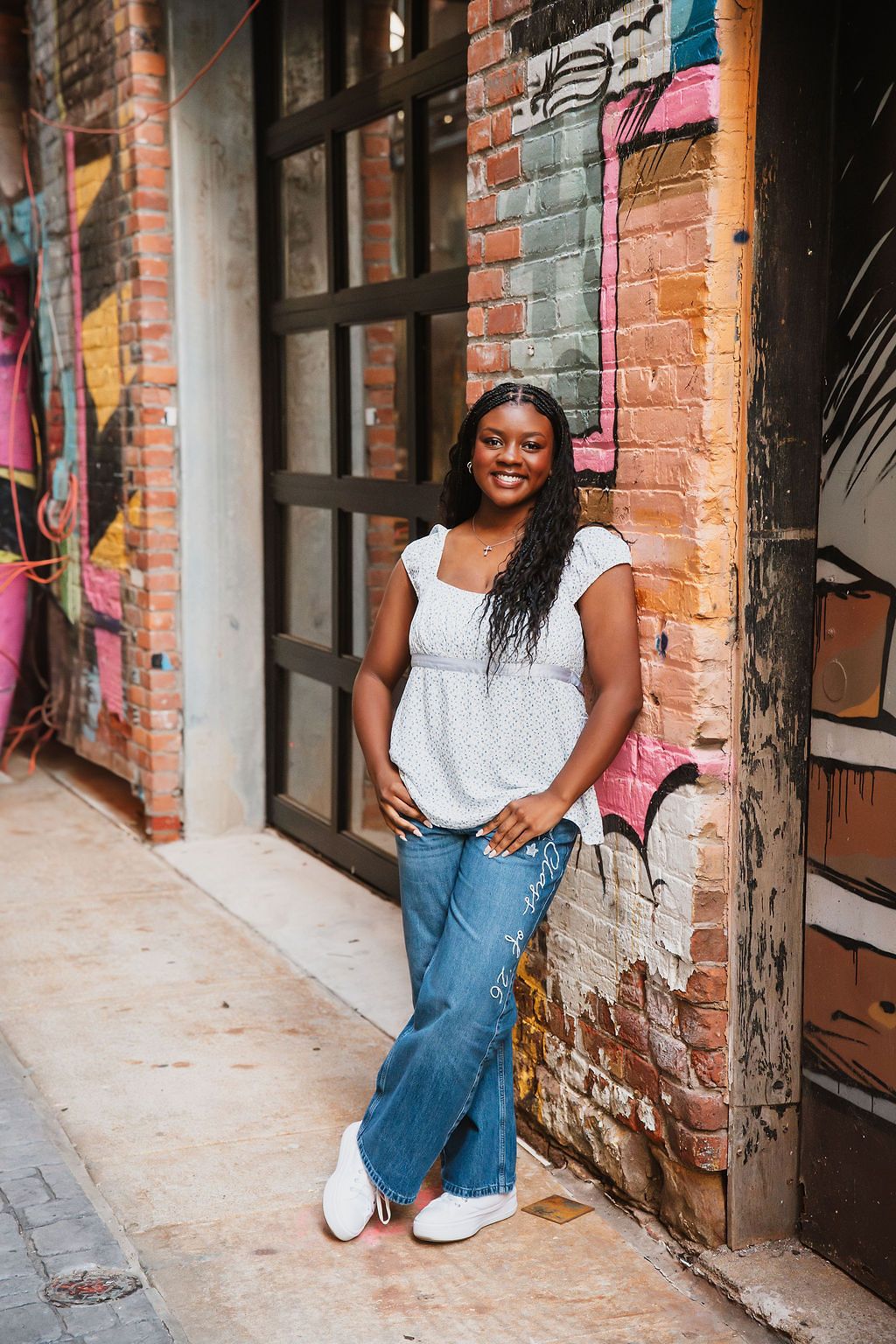 Smiling young Black woman in white top and jeans leaning against a colorful graffiti brick wall outdoors.