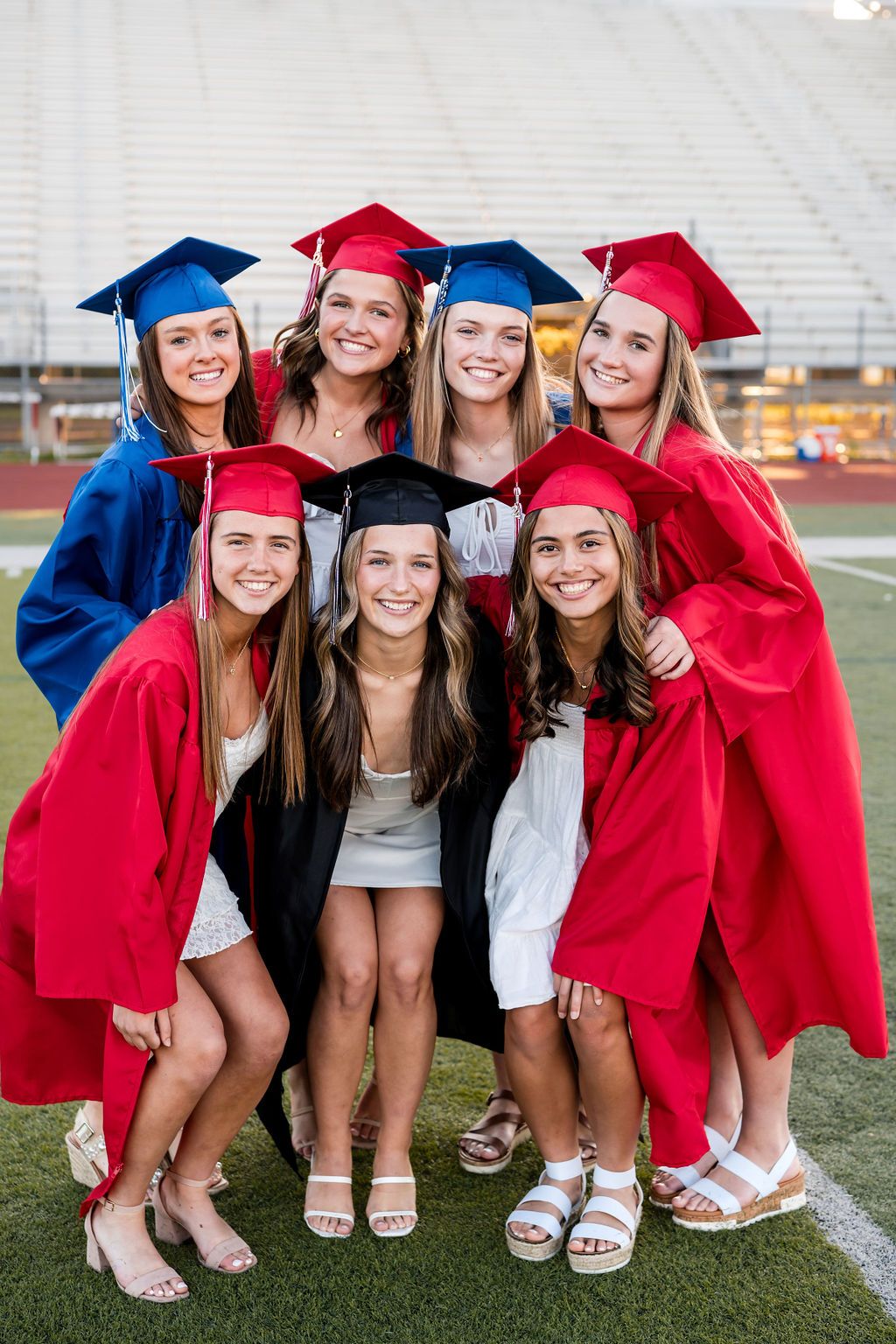 A group of graduates wearing red and blue caps and gowns pose together on a football field during graduation.