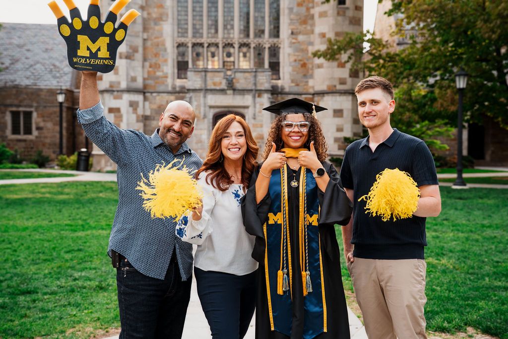 Graduates in black caps and gowns celebrate with yellow pom poms on a university campus lawn in autumn.
