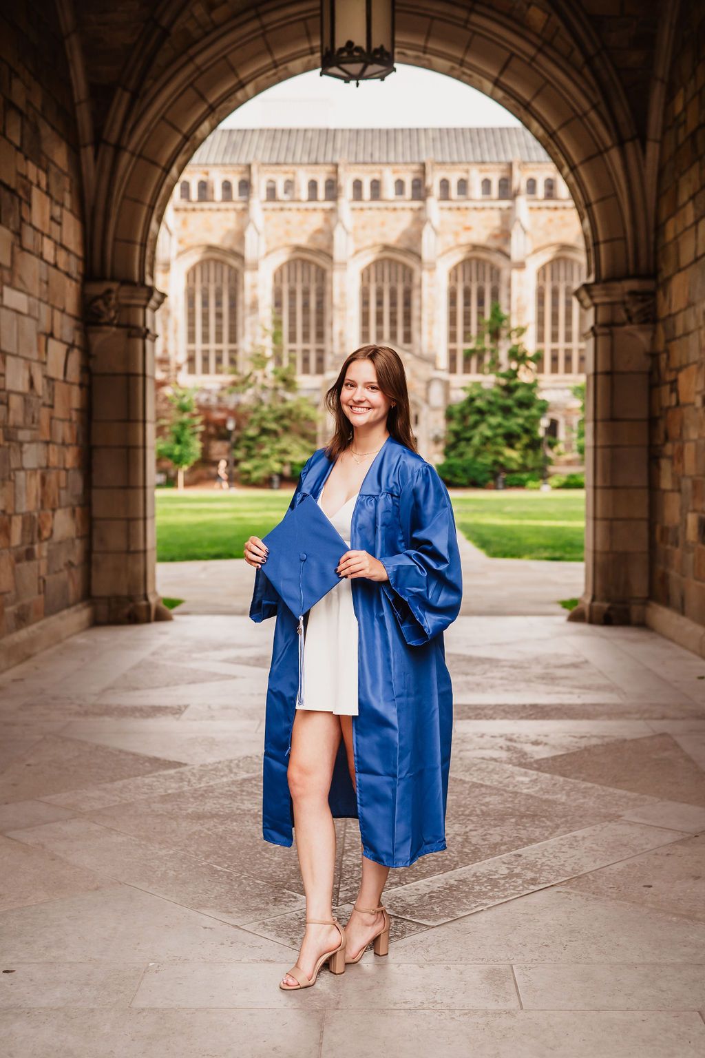 Graduate in blue gown stands under ornate stone archway with gothic university buildings in background.