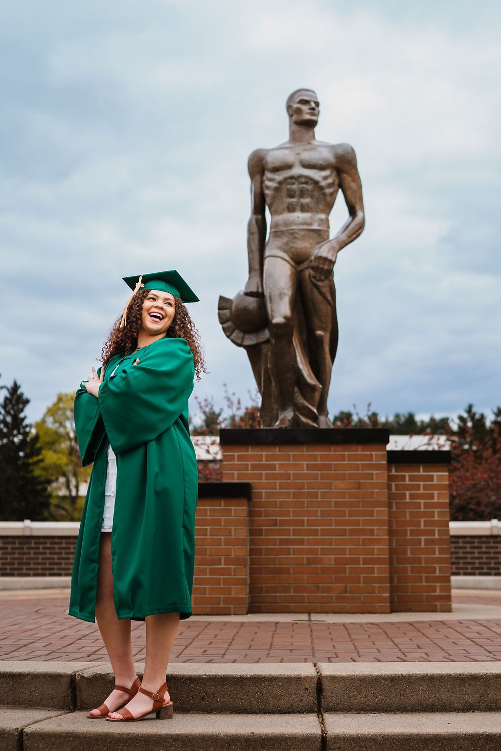 Graduate in green cap and gown poses playfully near bronze statue on brick wall during outdoor graduation photoshoot.