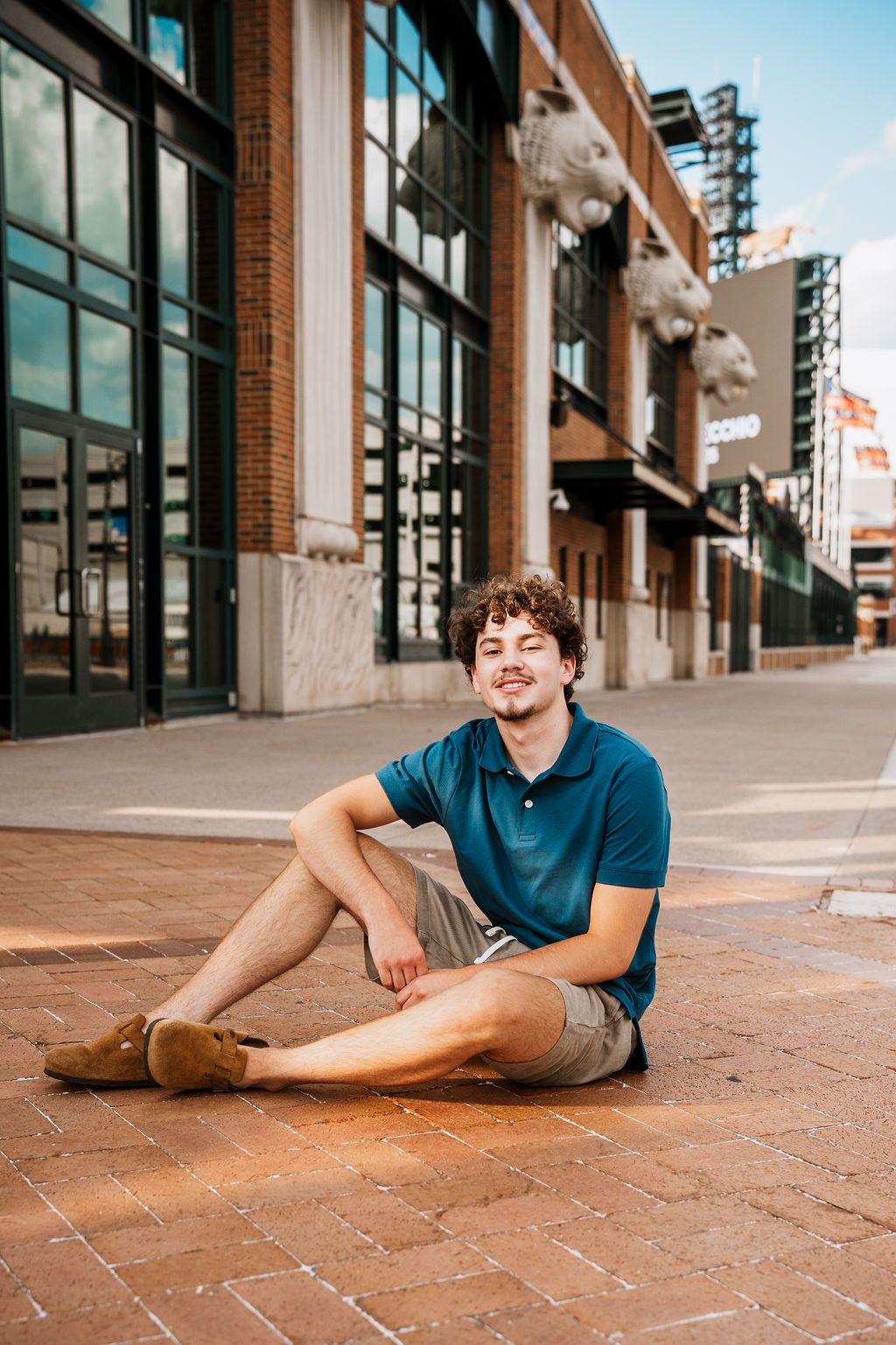 Someone sits casually on brick pavement wearing a blue polo shirt and khaki shorts outside a modern building facade.