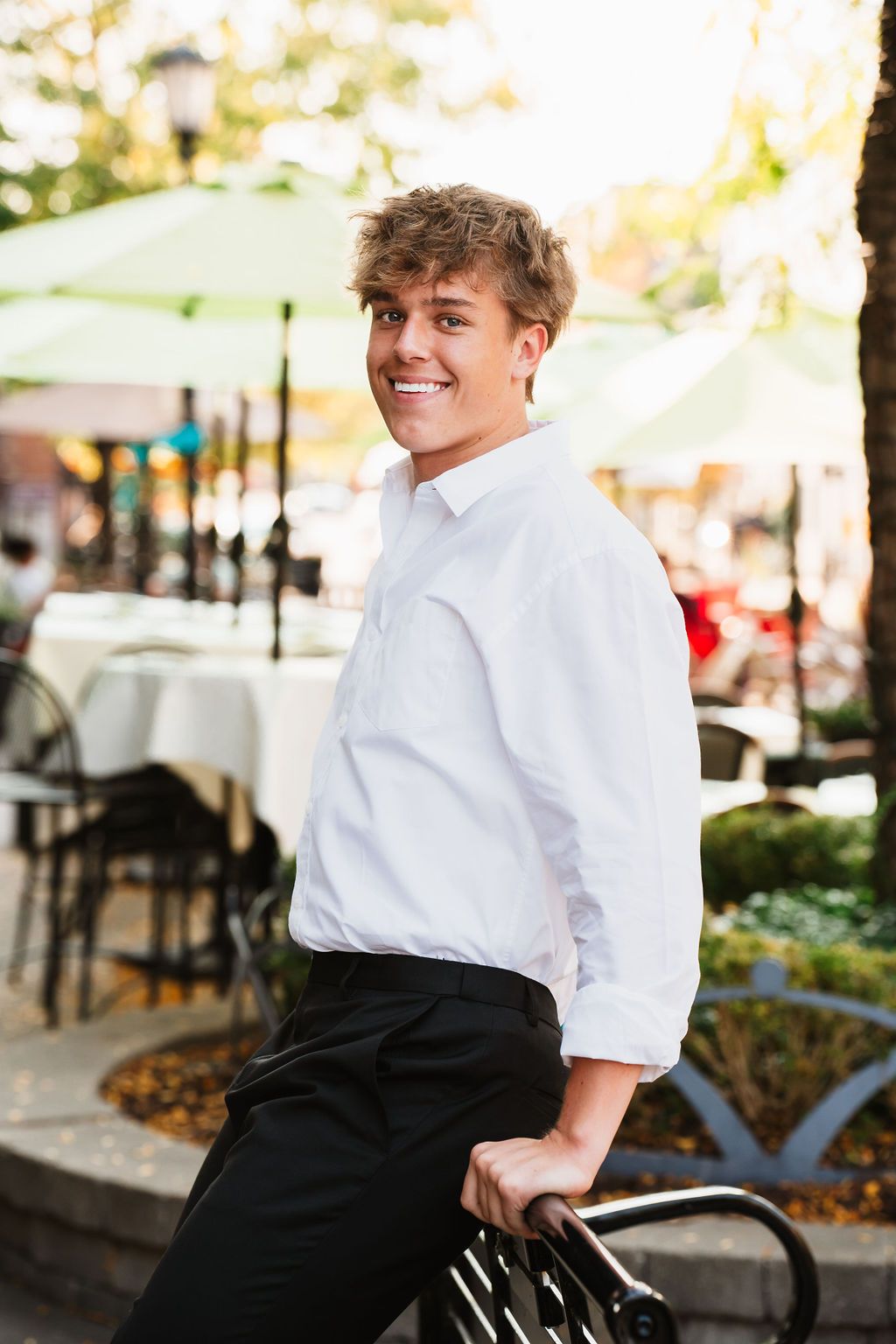 Smiling young man in white shirt and black pants leaning on outdoor cafe railing on a sunny day.