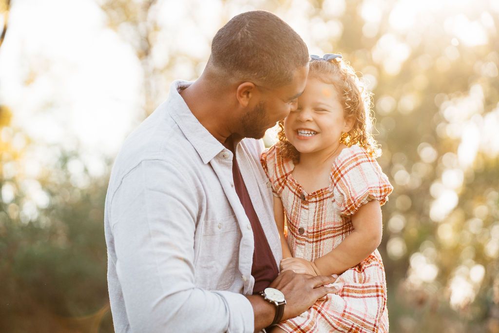 A father holds his laughing young daughter outdoors in warm golden sunlight, sharing a joyful moment together.