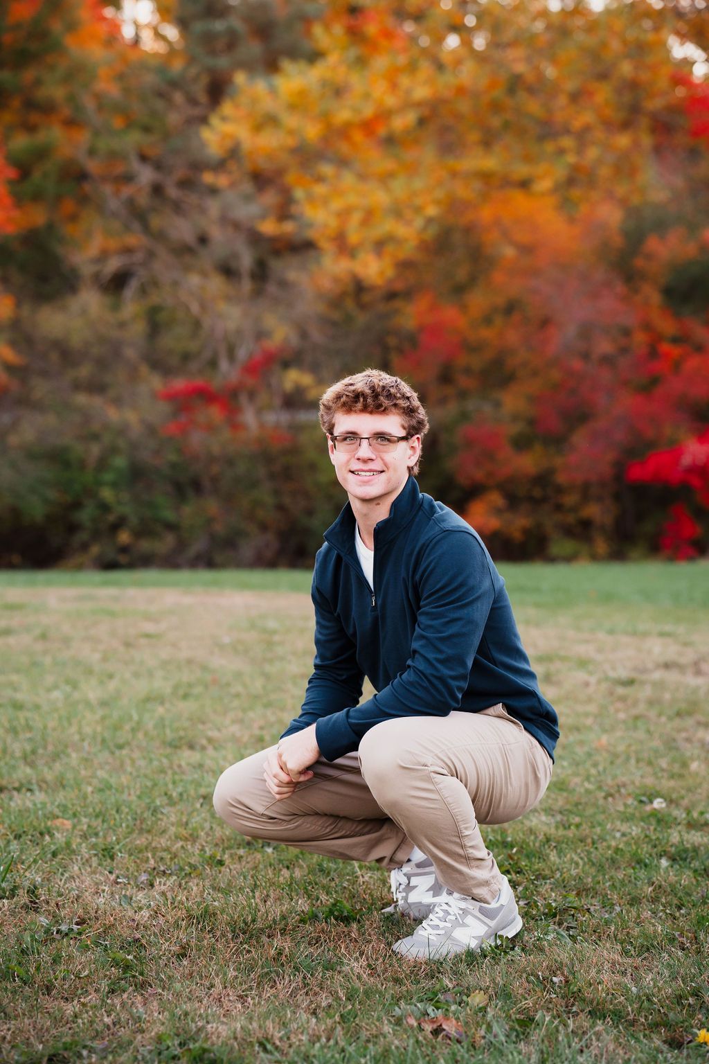 Young man in navy jacket and khaki pants crouching on grass with vibrant autumn foliage in background.