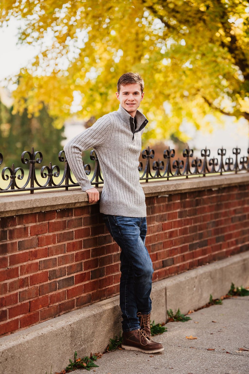 Young man in gray sweater and jeans leaning against brick wall with ornate iron fence in autumn setting.
