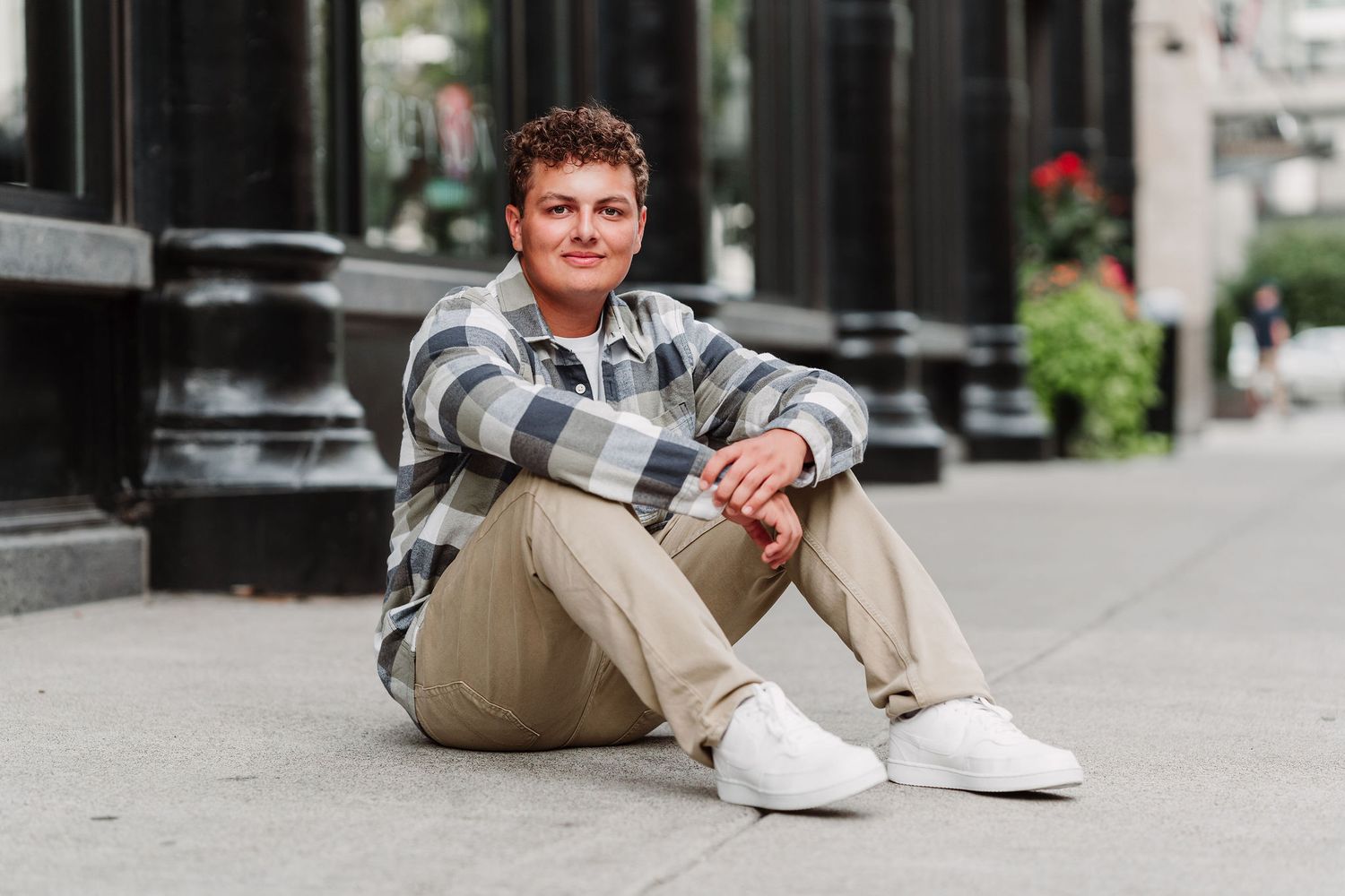 Young man in plaid shirt and khakis sitting relaxed on urban sidewalk, smiling confidently in casual outdoor portrait.