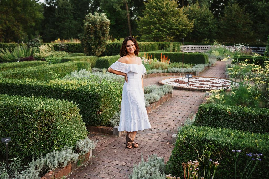 A person in a white off-shoulder dress stands on a brick path surrounded by manicured garden hedges and flowers.
