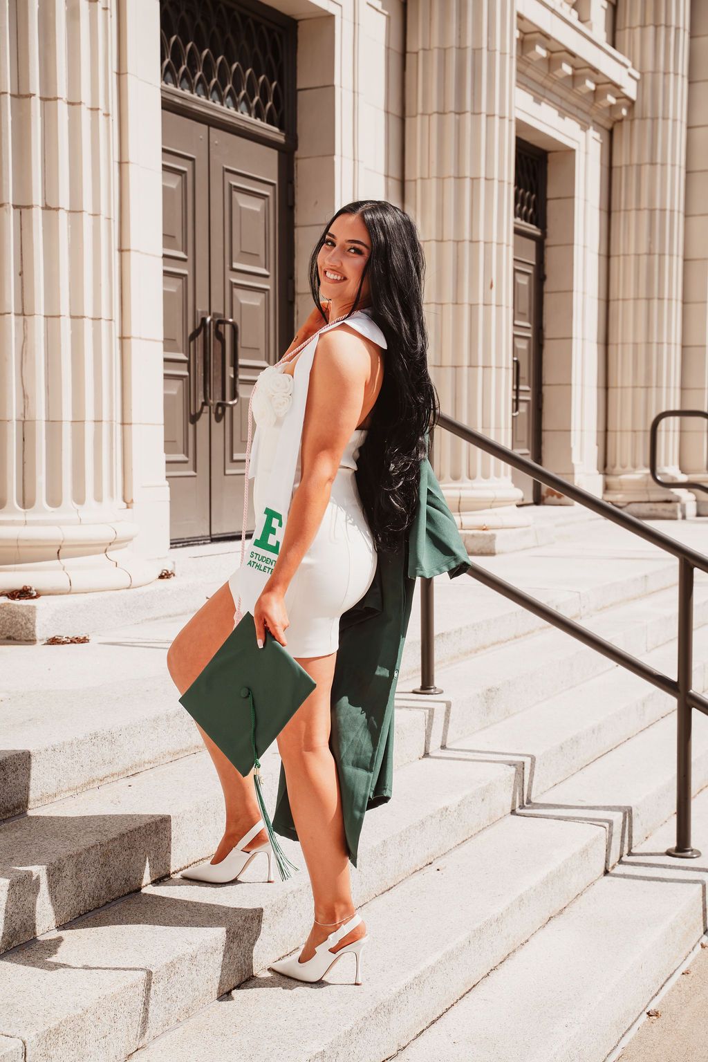 Smiling graduate in white dress holding green cap and gown on university steps in bright sunlight.
