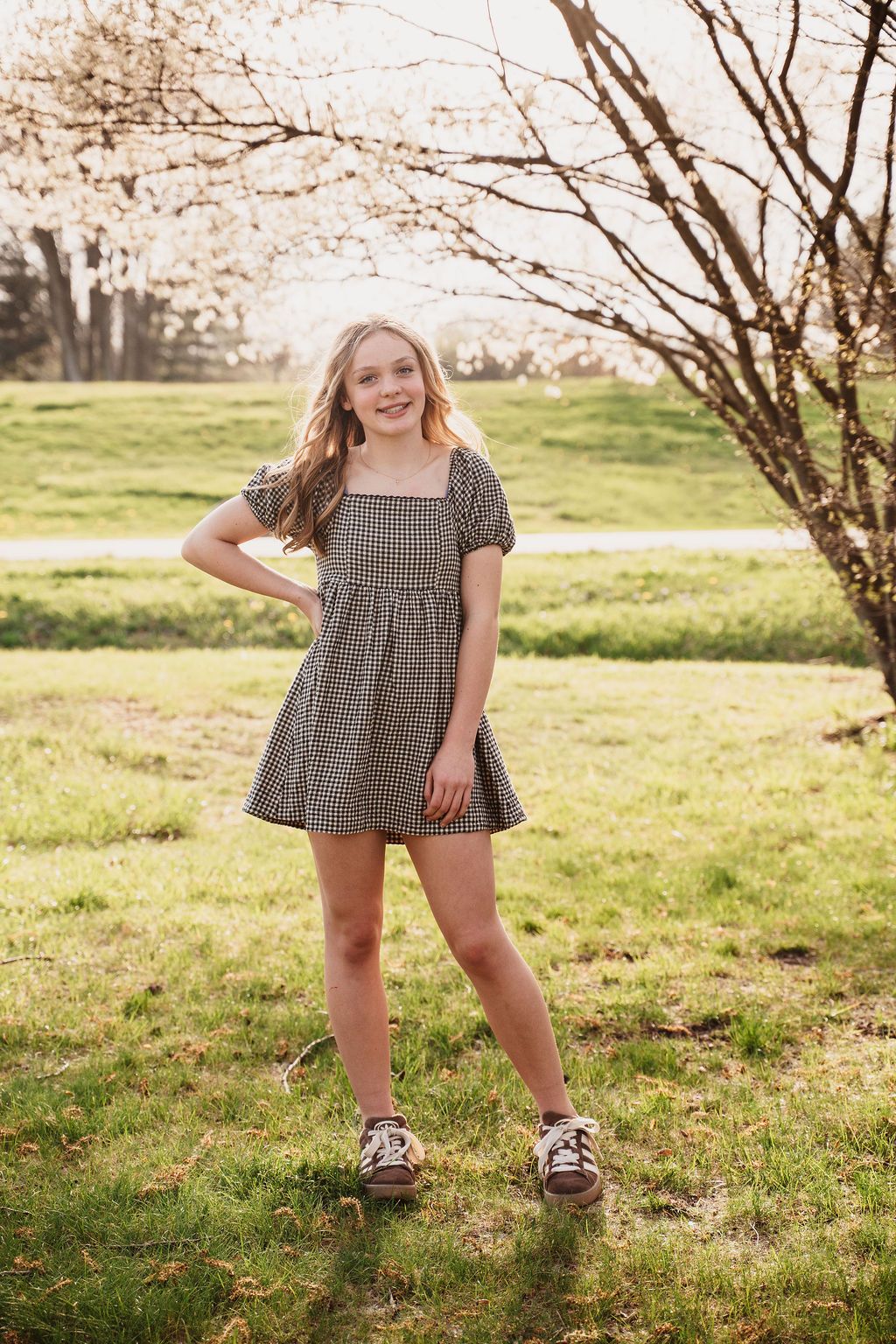 A smiling young girl in a gingham dress poses outdoors near a flowering tree on a sunny spring day.