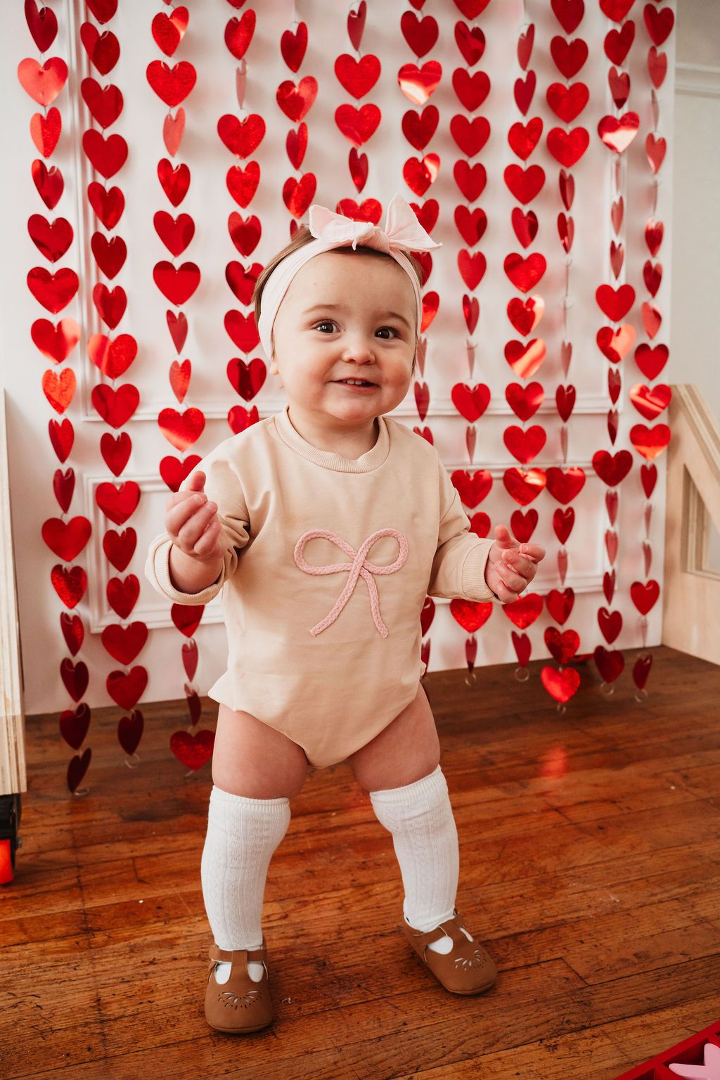 Smiling baby girl in beige onesie and white knee socks stands before red heart garland backdrop.