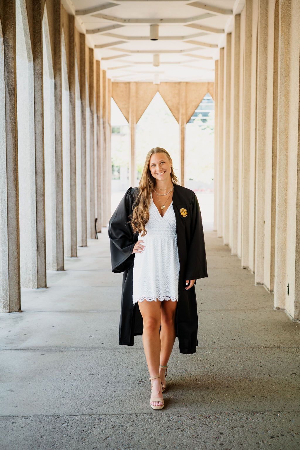 Graduate poses in white dress and black blazer under architectural colonnade with repeating pillars.