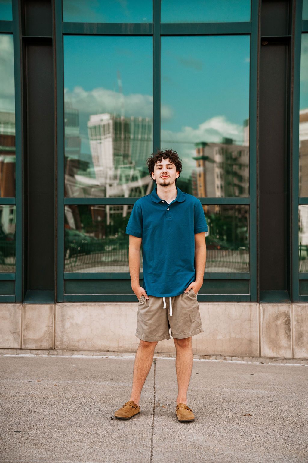 Person in a blue polo shirt and khaki shorts stands on concrete sidewalk with city buildings in background.