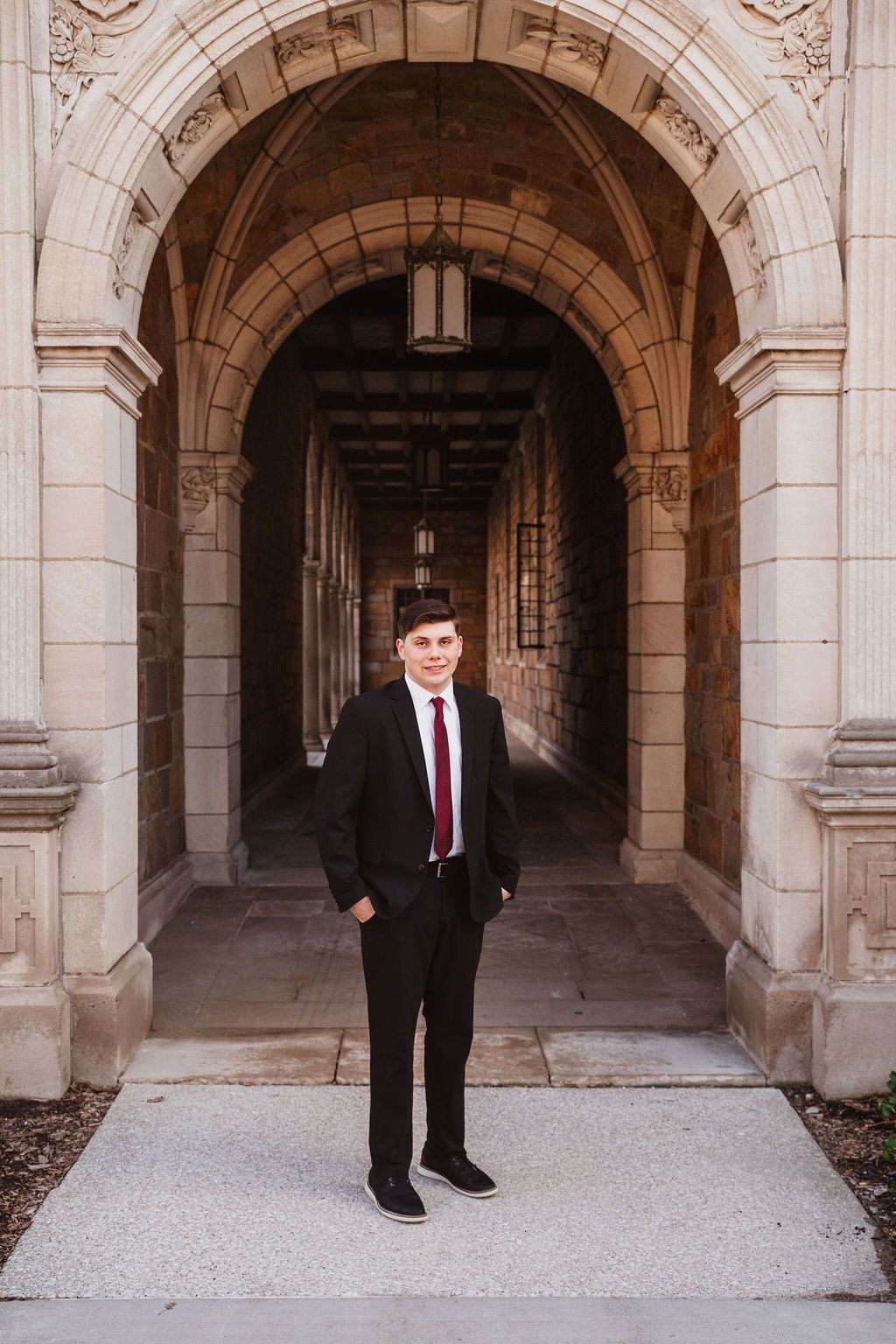 Young man in black suit and red tie standing confidently under ornate stone archway corridor.