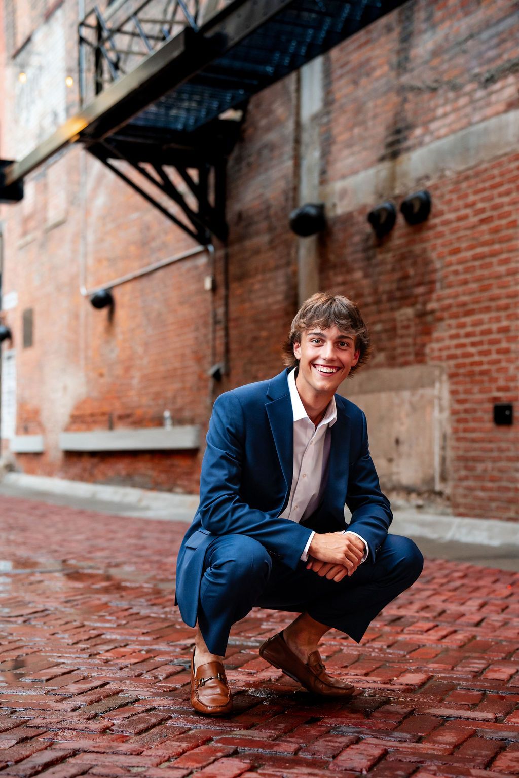 Young man in blue suit crouching on red brick alleyway, smiling confidently in urban setting.
