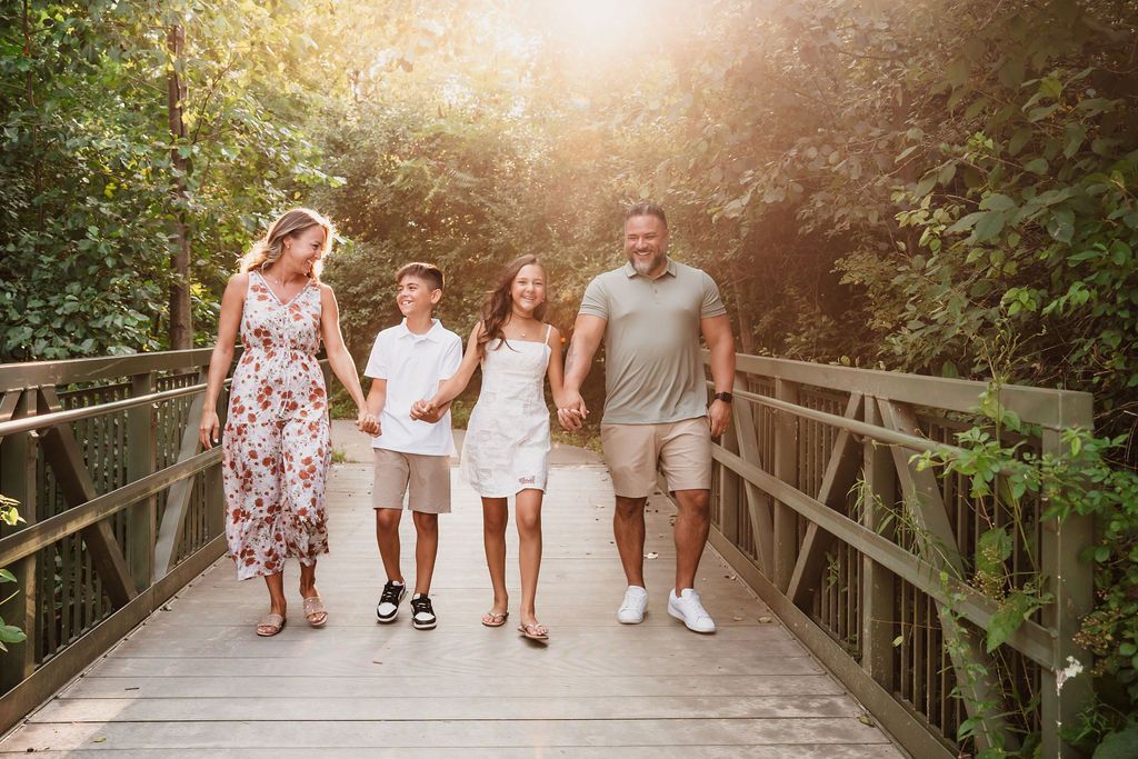 Family of four walking hand-in-hand on a wooden bridge surrounded by lush green trees in warm sunlight.
