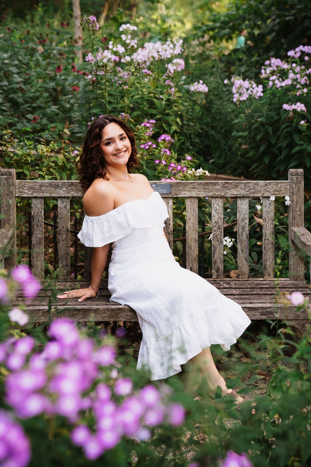 A white dress contrasts beautifully against a garden bench surrounded by blooming purple flowers and lush greenery.