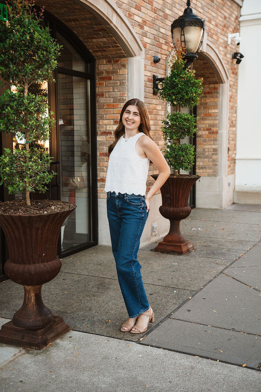Smiling young woman in white top and jeans poses outside brick building with potted shrubs.