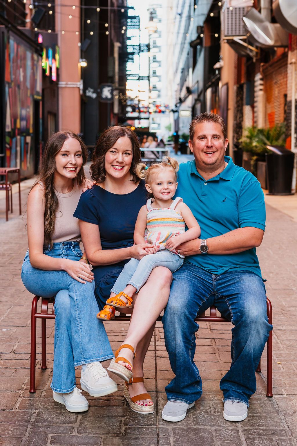 Happy family of four posing together on a bench in a charming outdoor alley with string lights.