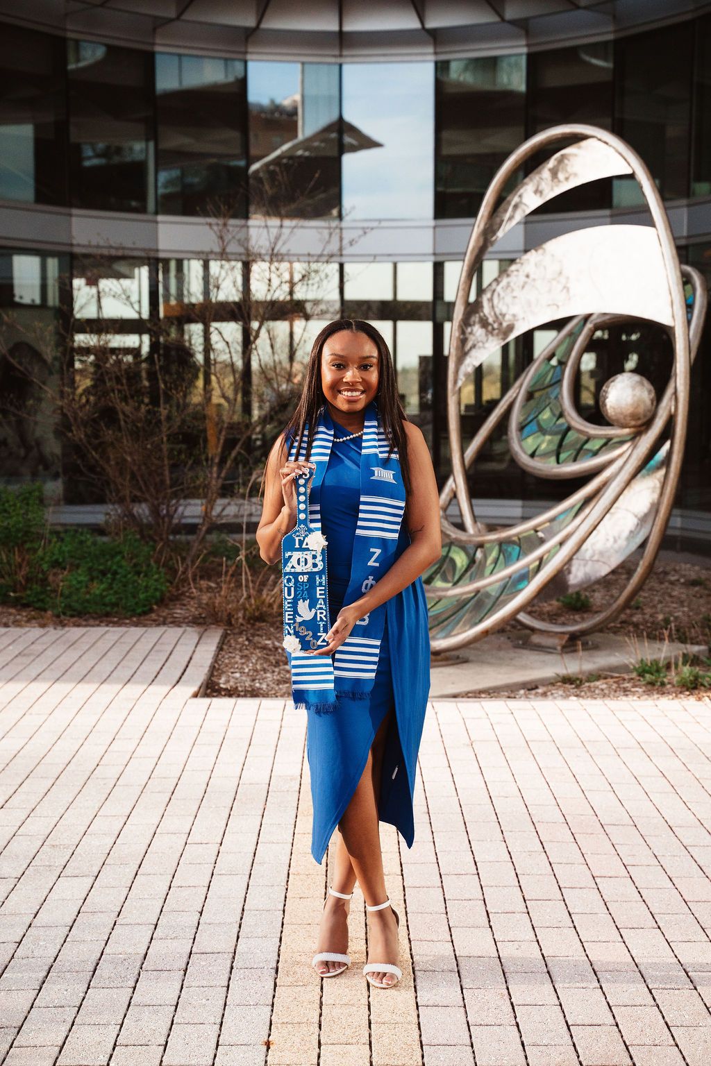 Smiling young woman in blue dress and graduation stole poses outdoors near modern sculpture at university campus.
