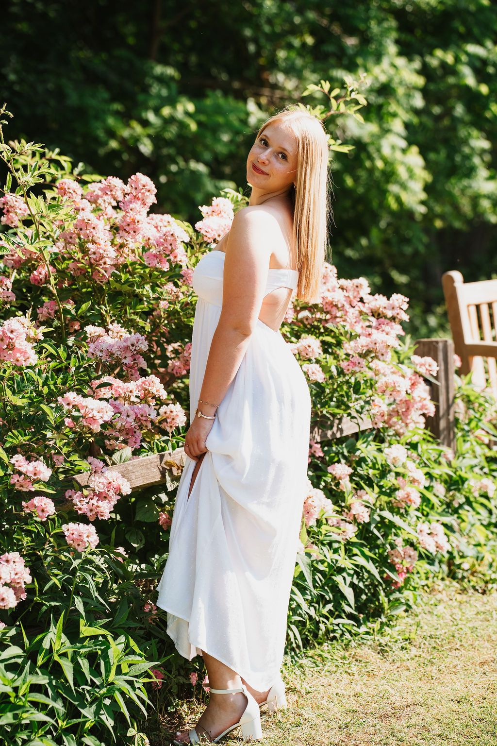 Young blonde woman in white dress posing beside pink flowering bushes in a sunny garden.