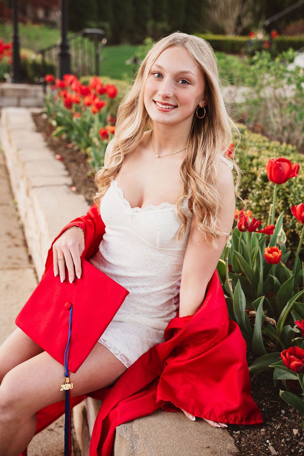 Smiling blonde graduate in white dress with red cap and gown sitting among red tulips outdoors.