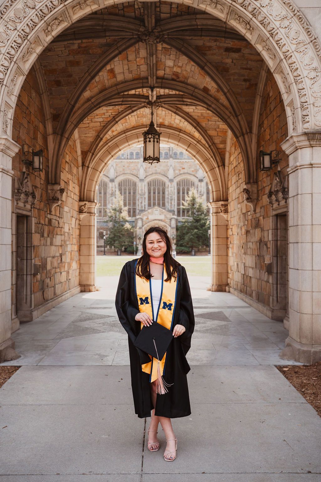 University of Michigan graduate smiling in cap and gown under Gothic stone archway on campus.