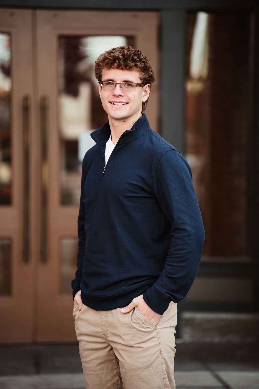 Young man in navy quarter-zip and khakis smiling outdoors near wooden doors.