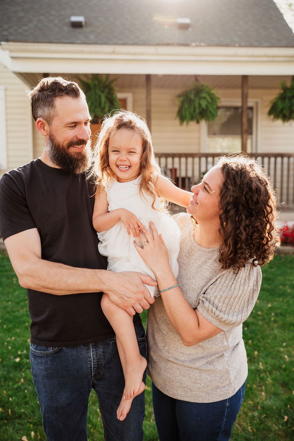 Happy family with bearded dad, curly-haired mom, and laughing little girl posing in front of a house.