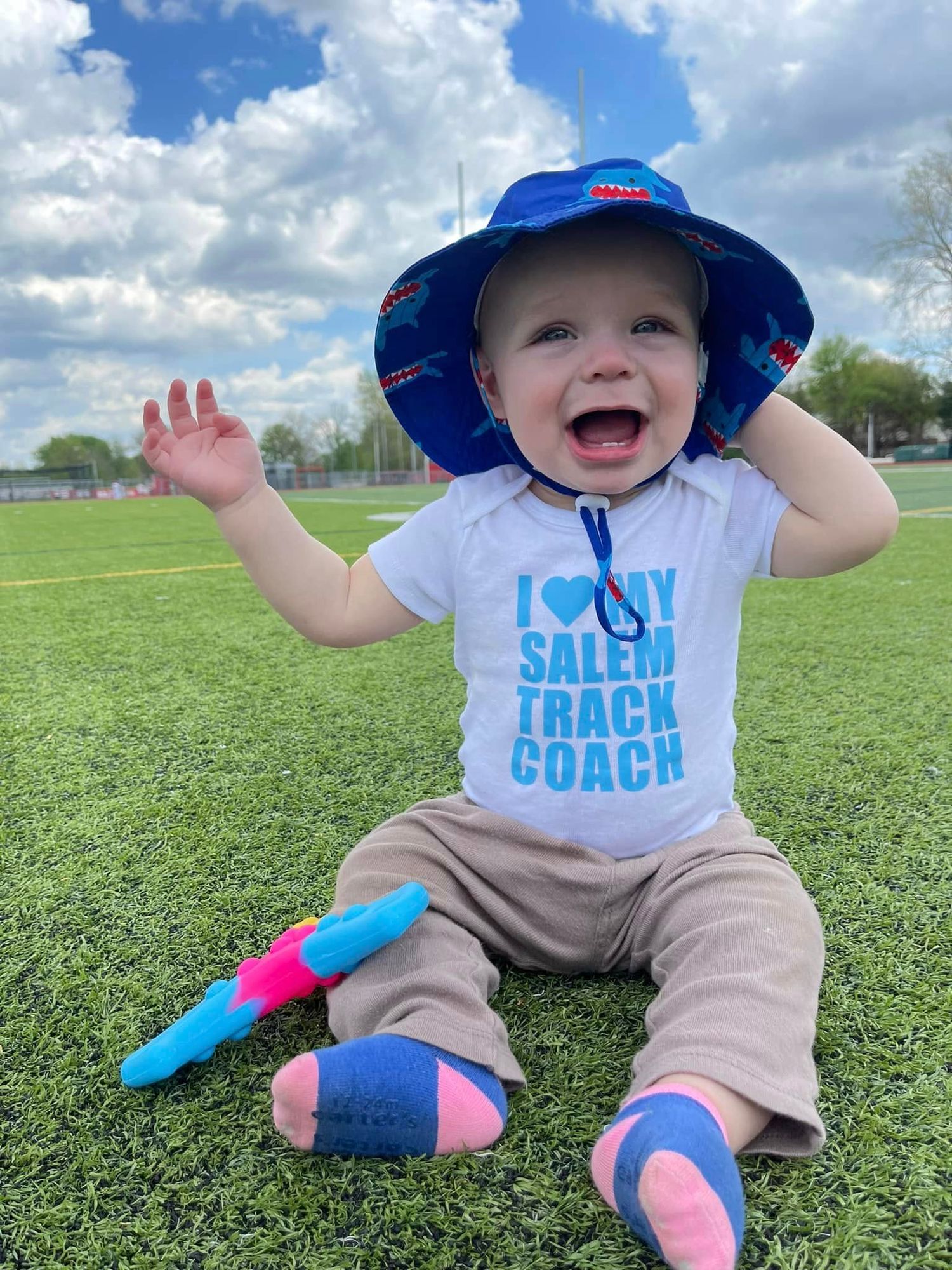 Happy baby wearing a blue sun hat and Salem Track Coach onesie sitting on green turf field.