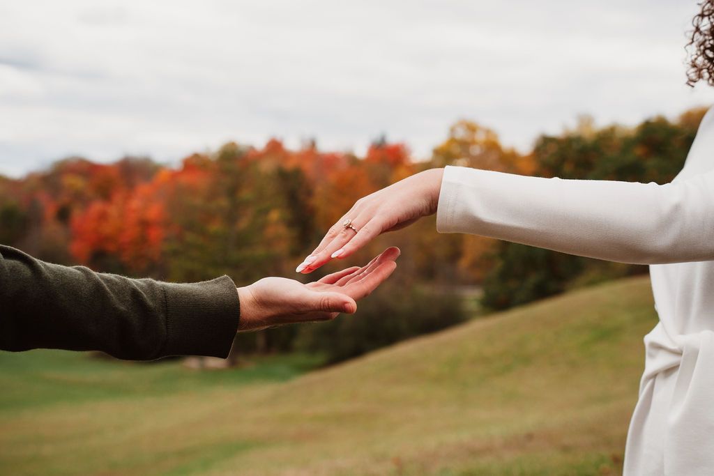 Two people reaching toward each other outdoors in autumn, with colorful fall foliage in the background.