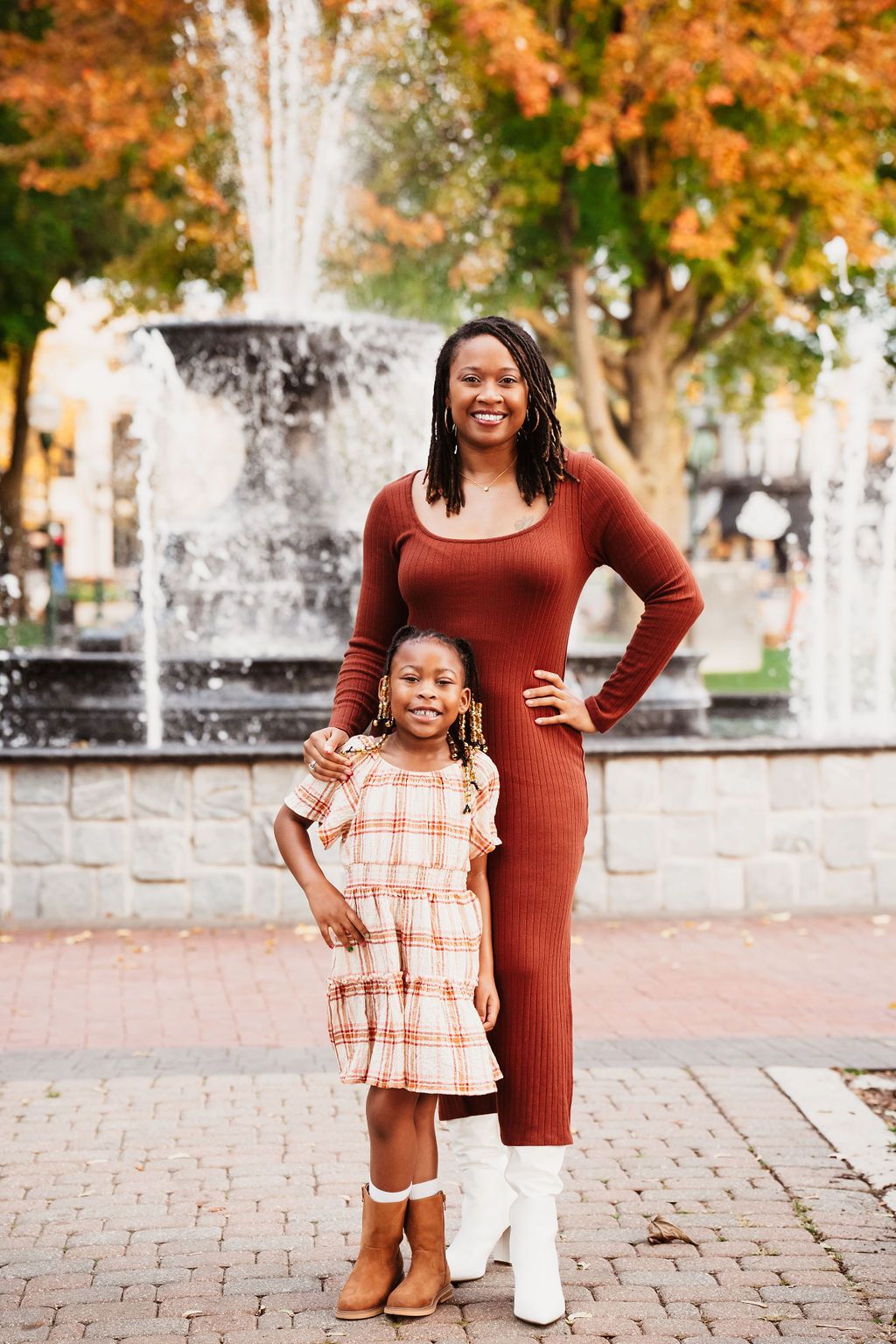 A woman and young girl in matching rust-toned outfits pose together by a fountain in an autumn park.