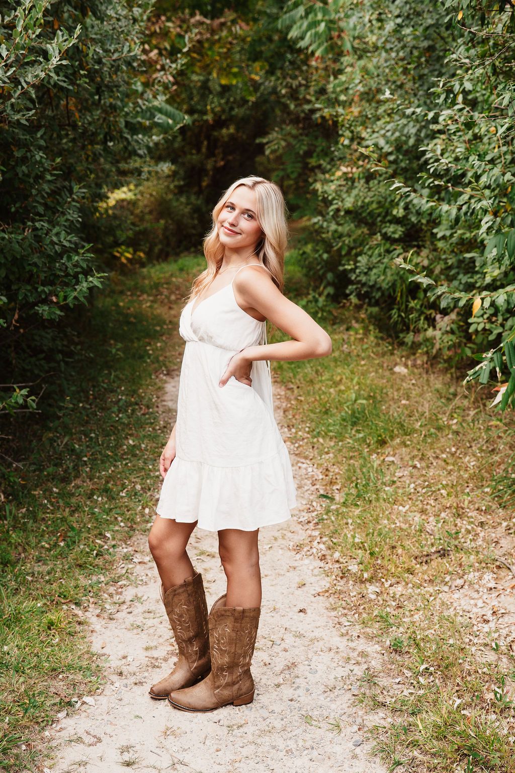 Young blonde woman in white sundress and brown cowboy boots posing on a dirt path surrounded by lush green trees.
