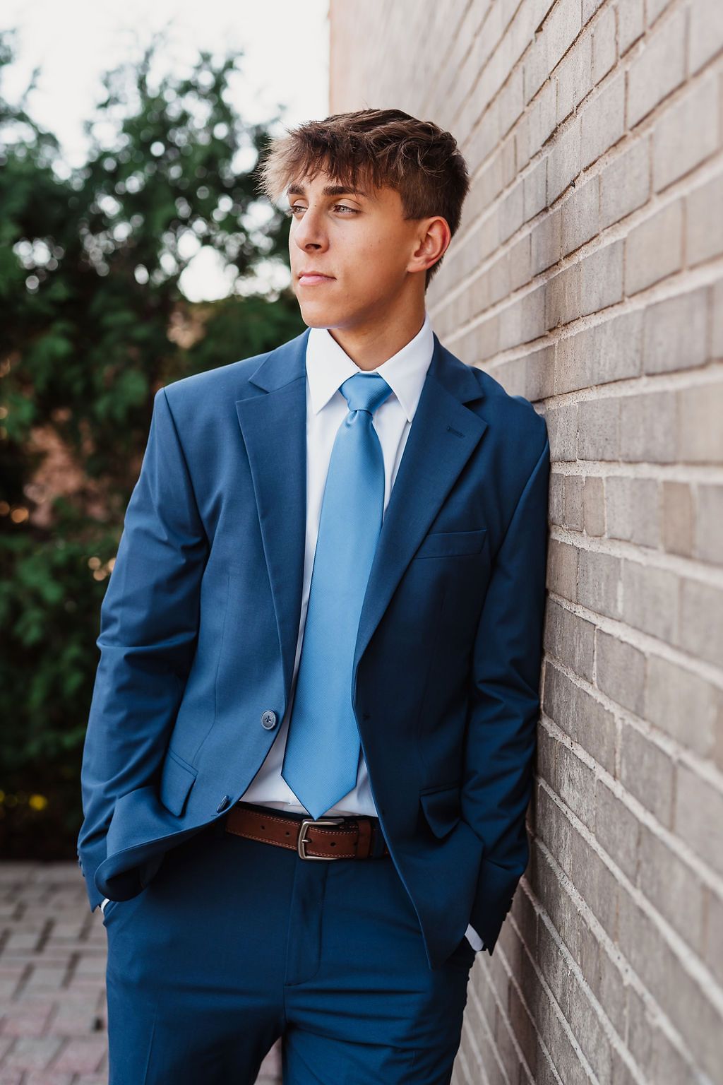 Young man in navy blue suit and light blue tie leaning against brick wall outdoors.