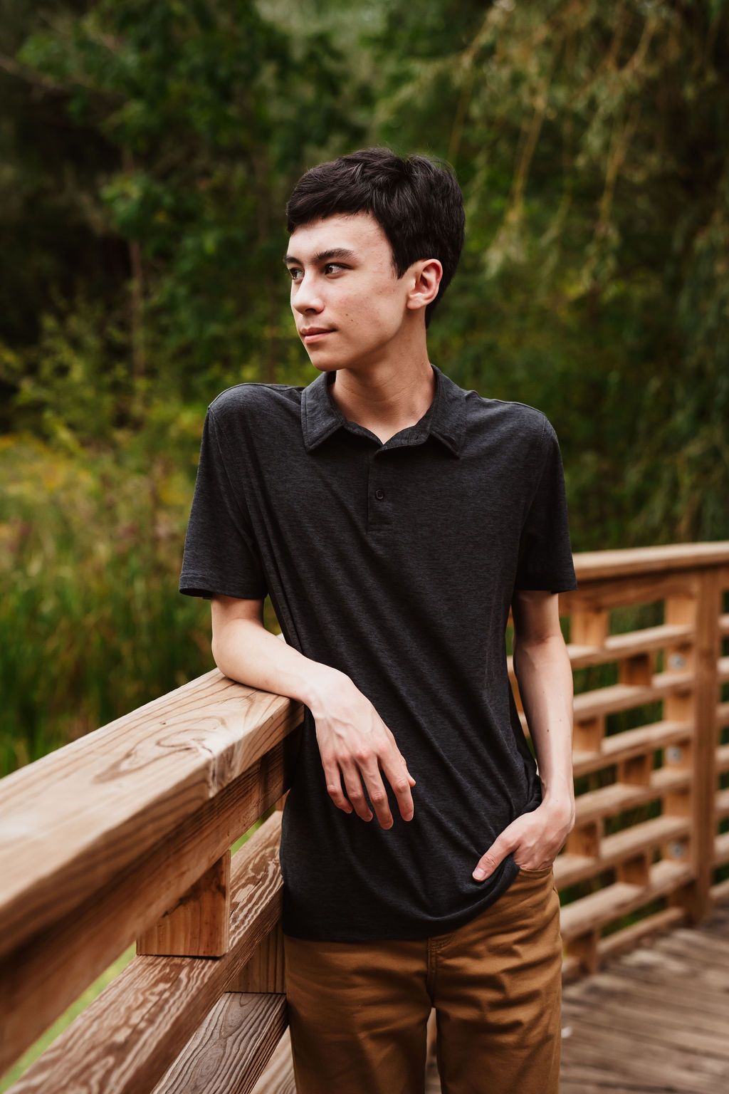 Young man in black polo shirt leaning on wooden bridge railing surrounded by lush green nature.