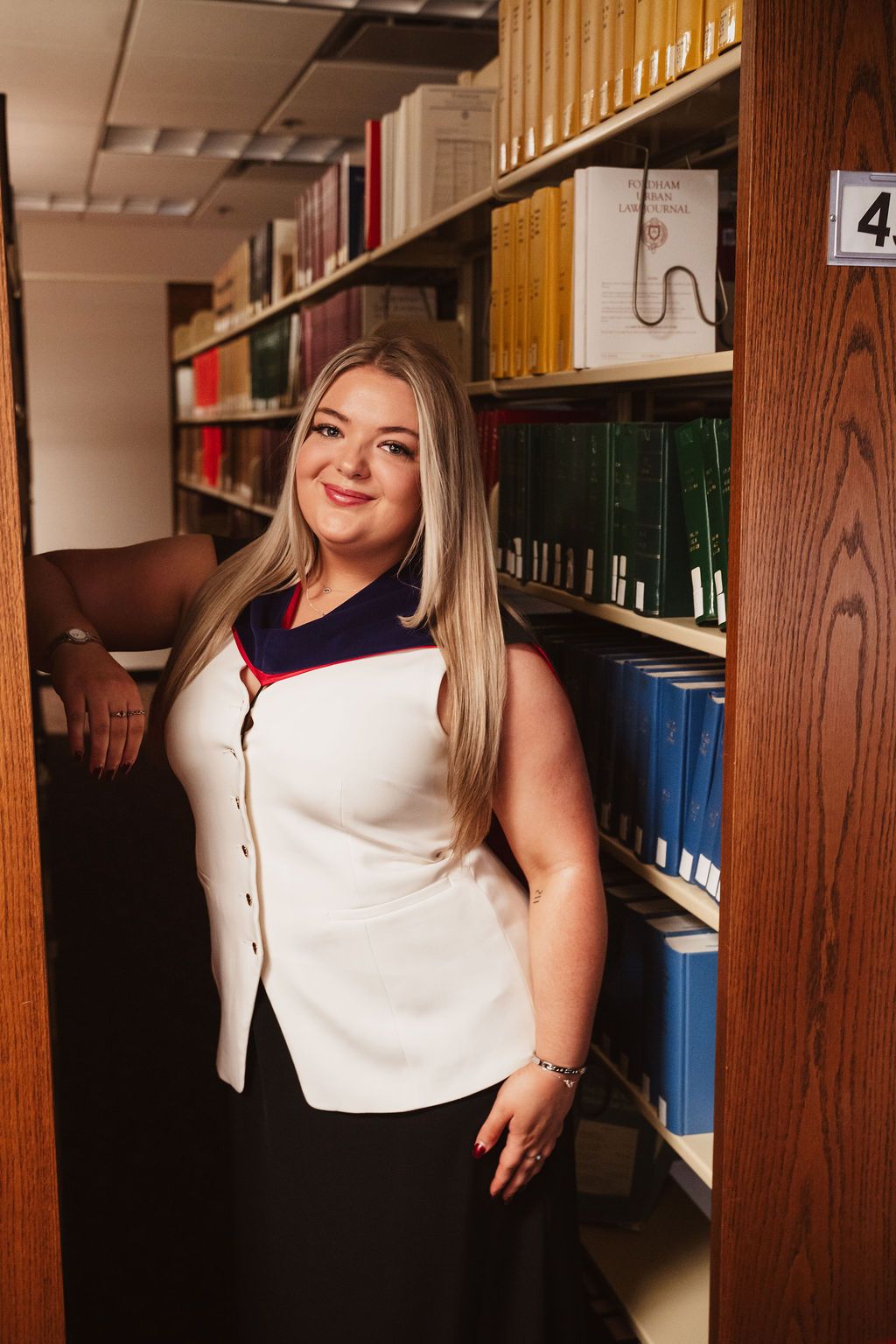 Young woman with blonde hair smiling in library aisle, wearing white vest top and black skirt, surrounded by bookshelves.