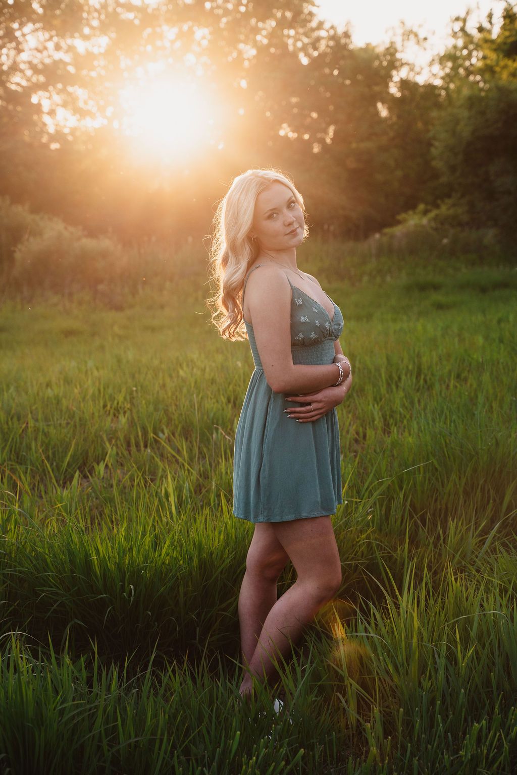 Blonde woman in teal dress standing in green field during golden sunset.