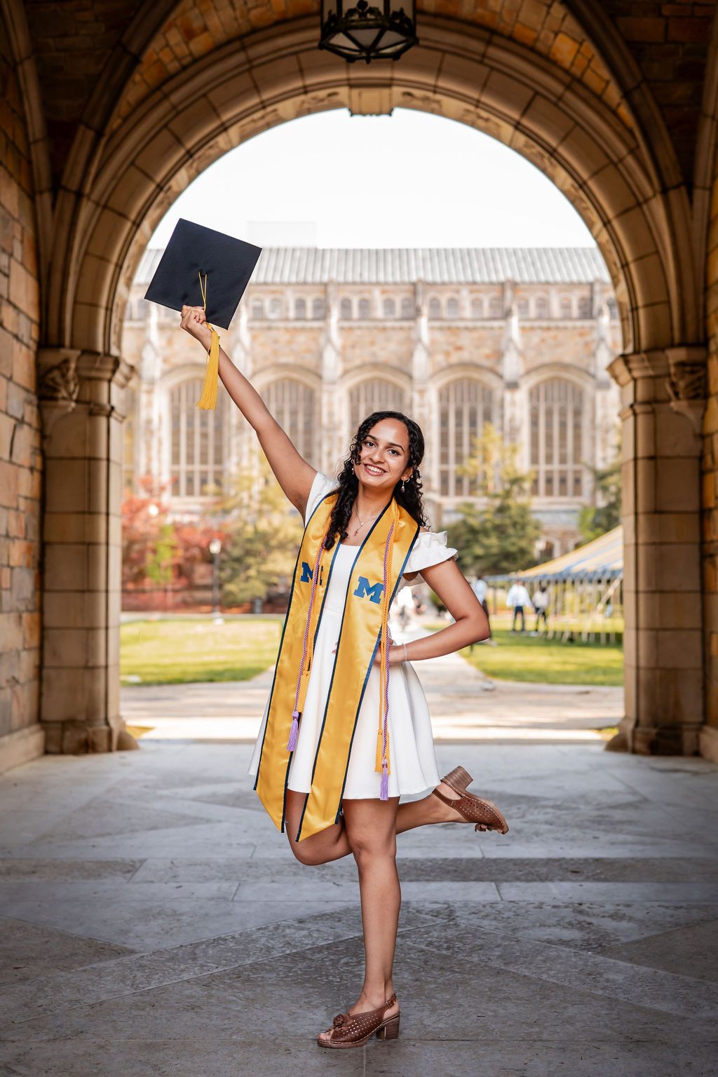 Graduate poses in gold stole under stone archway with gothic university architecture visible in background.