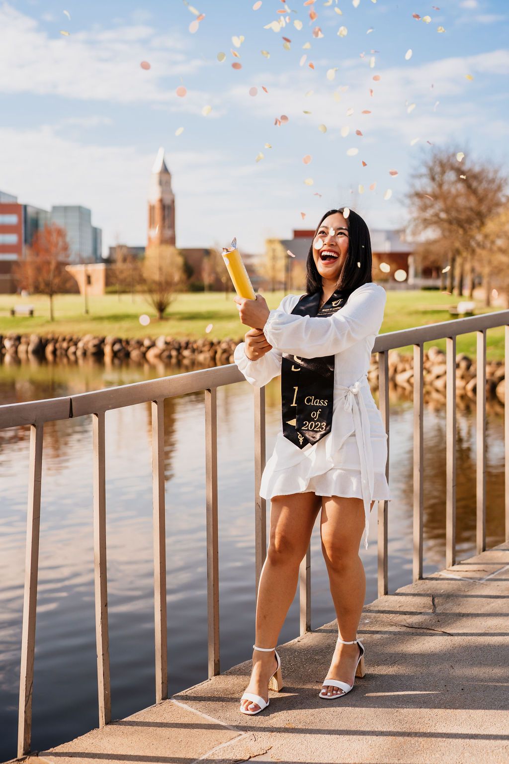 A graduation photoshoot on a sunny day by a riverside walkway with confetti floating through the air.