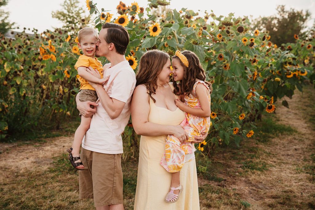 Family of four posing in a sunflower field, parents kissing children in golden light.