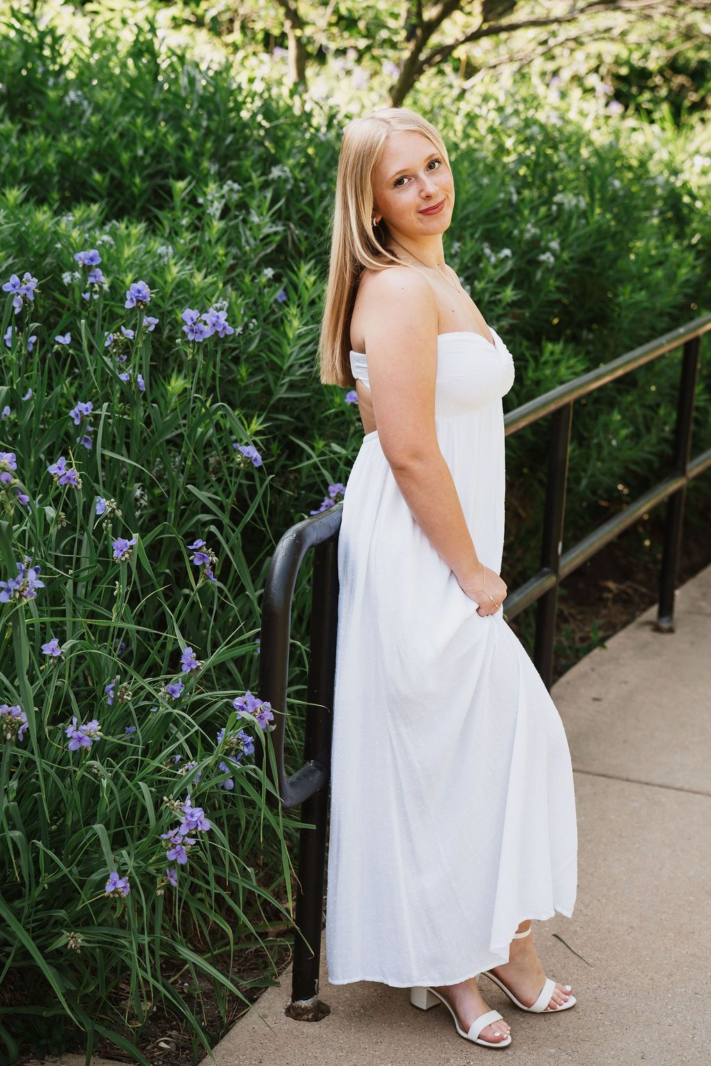 Blonde woman in elegant white maxi dress standing by railing with purple flowers and green garden background.