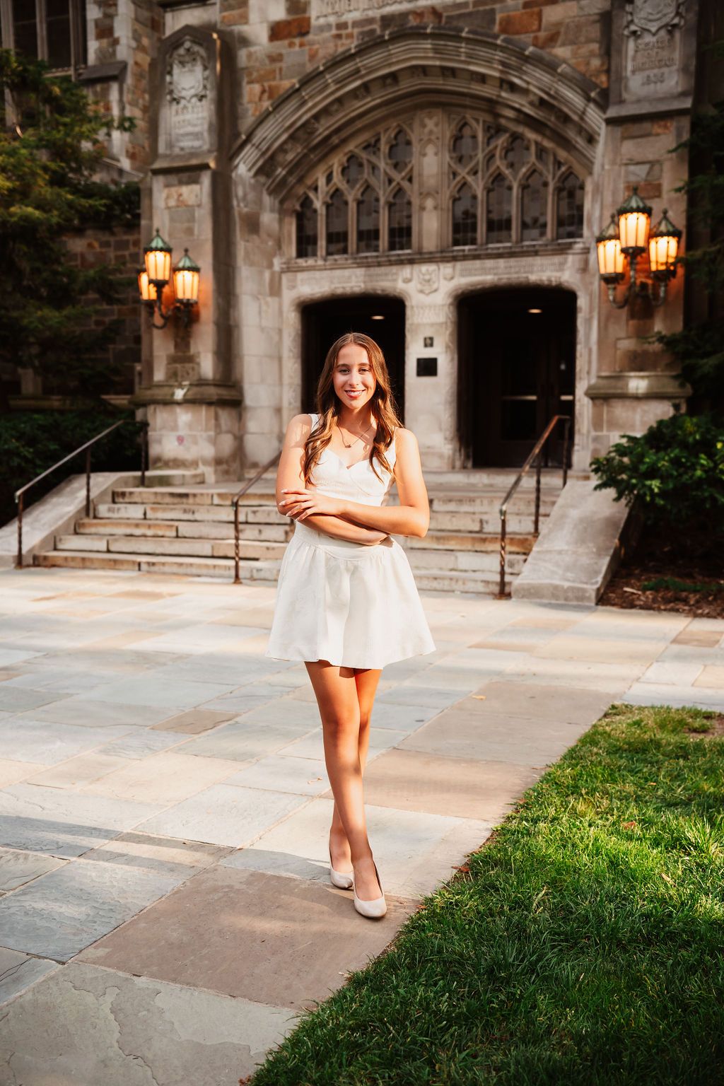 A student in a white dress poses on campus steps in front of a gothic stone archway with glowing lanterns.