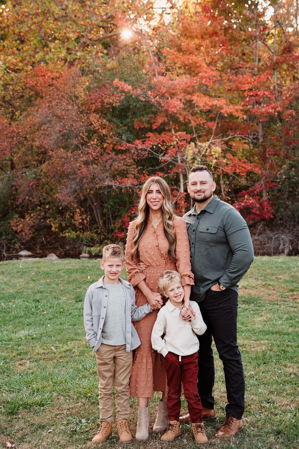 Family of four posing outdoors during fall season with vibrant red and orange foliage in background.