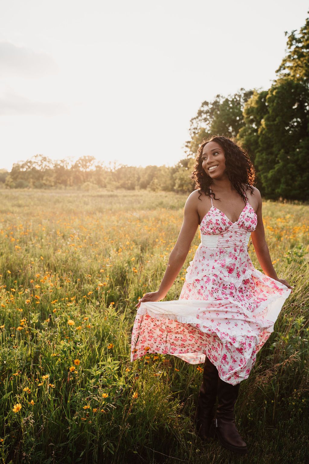 A smiling young Black woman in a floral sundress stands in a golden wildflower field at sunset.