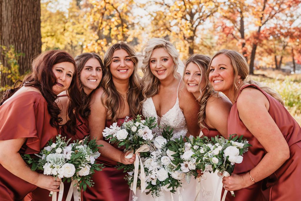 Bridesmaids in burgundy dresses hold white floral bouquets during outdoor autumn wedding.